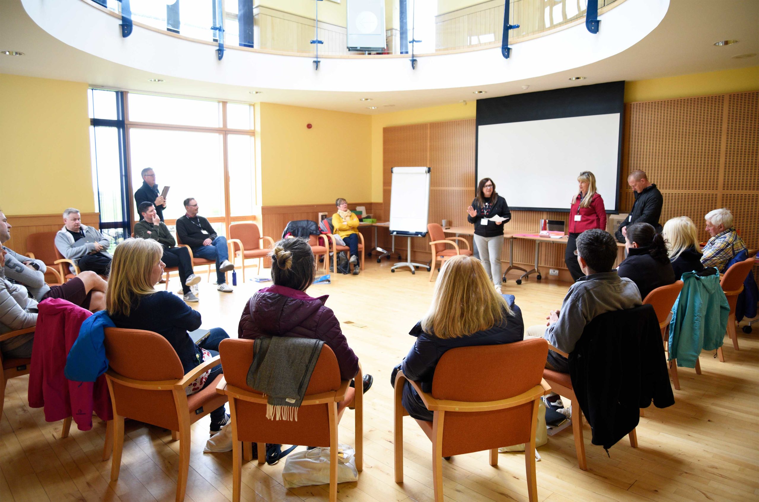 Group a giving presentation in front of a larger group sitting in chairs